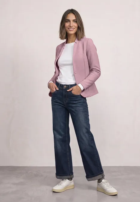 Eine Frau trägt ein rosa Sakko, burgunderfarbenes Shirt, blaue Jeans und Loafers und hält eine braune Clutch.