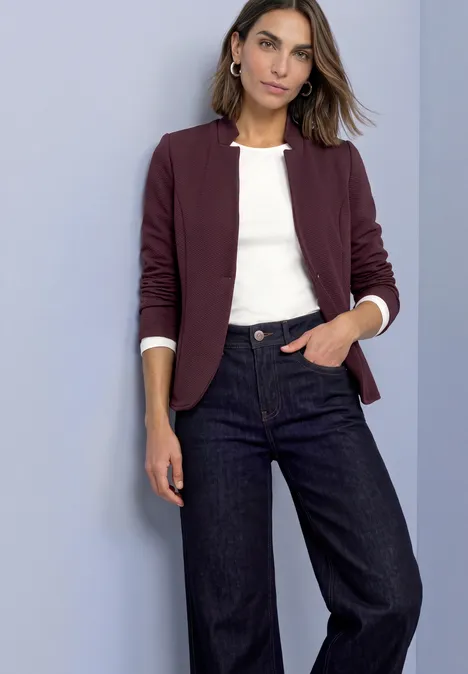Frau mit burgunderfarbenem Blazer, weißem Shirt und weit geschnittenen Jeans vor schlichtem Hintergrund.