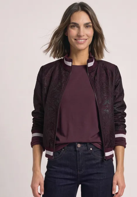 Blouson à motifs floraux berry maroon Porträt einer lächelnden Frau in lila Bomberjacke und dunkler Jeans vor hellem Hintergrund.