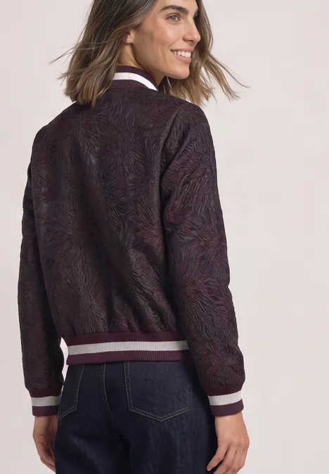 Blouson à motifs floraux berry maroon Frau von hinten in dunkelroter, gemusterter Bomberjacke mit weißem Kragen und Bündchen, dazu Jeans.