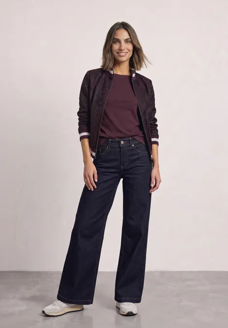 Blouson à motifs floraux berry maroon Frau in bordeauxroter Bluse, violetter Bomberjacke, weiten Jeans und weißen Sneakern.