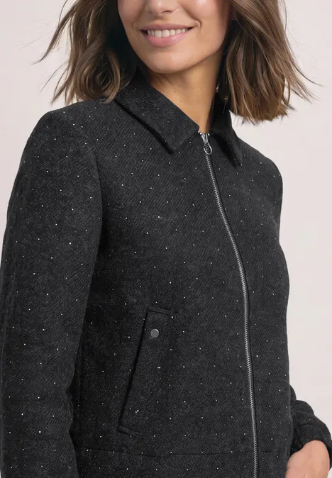 Eine Frau mit braunen Haaren trägt eine schwarze, strukturierte Jacke mit Reißverschluss und Stehkragen.