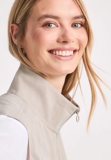 Eine lächelnde Frau mit blondem Haar trägt eine helle Weste vor weißem Hintergrund.