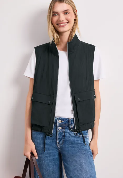 Vrouw draagt zwart gewatteerd vest op wit T-shirt, blauwe jeans en houdt een handtas.