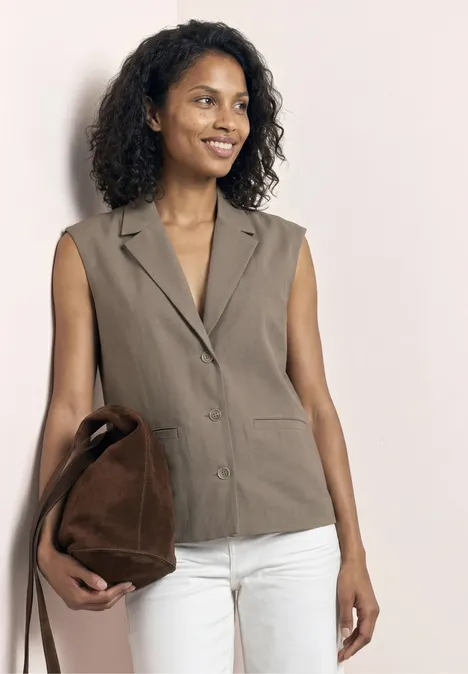Vrouw met mouwloos taupecolbert, witte broek en bruine tas.