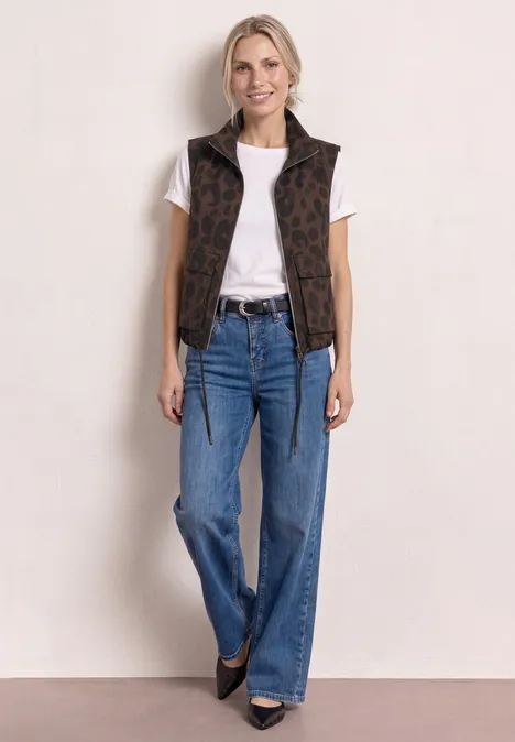 Frau in weißem T-Shirt, braunem Muster-Ärmelloser Weste, blauen Schlagjeans und schwarzen Schuhen.