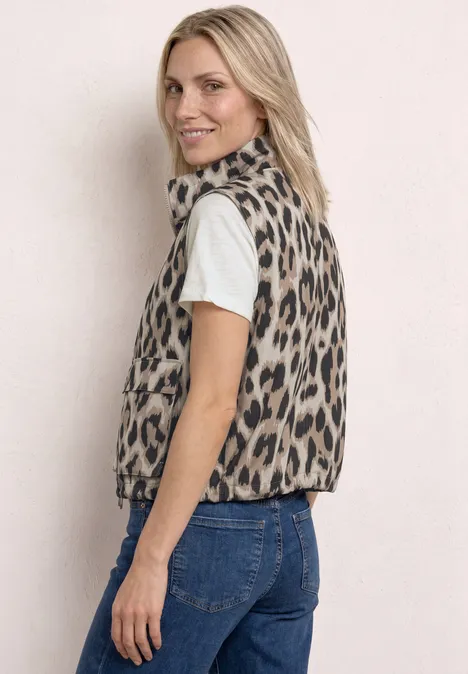 Frau seitlich in Jeans, weißem T-Shirt und Leopardenfell-Weste vor heller Wand