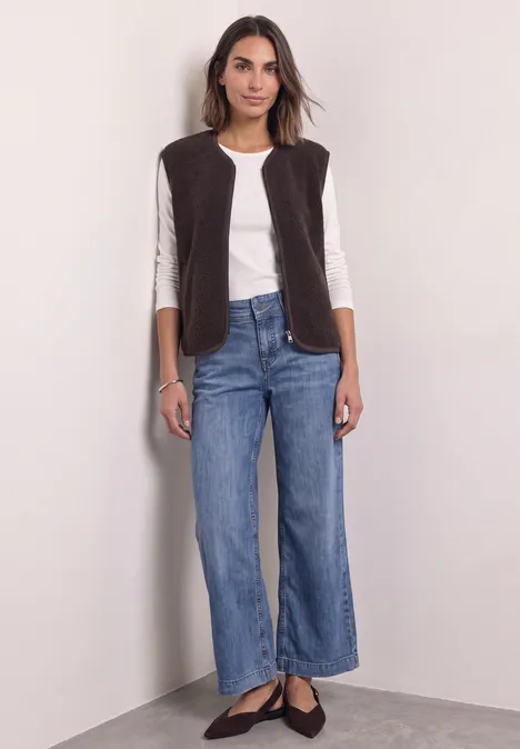 Eine Frau vor heller Wand trägt weiße Bluse, braune Weste, weite Jeans und schwarze Ballerinas.