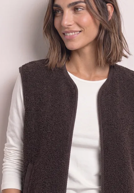 Eine lächelnde Frau trägt einen braunen Teddy-Fleece-Weste über einem weißen Langarmshirt.