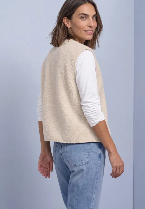 Frau schaut über die Schulter und trägt eine beige Fleeceweste über weißem Langarmshirt und Jeans.