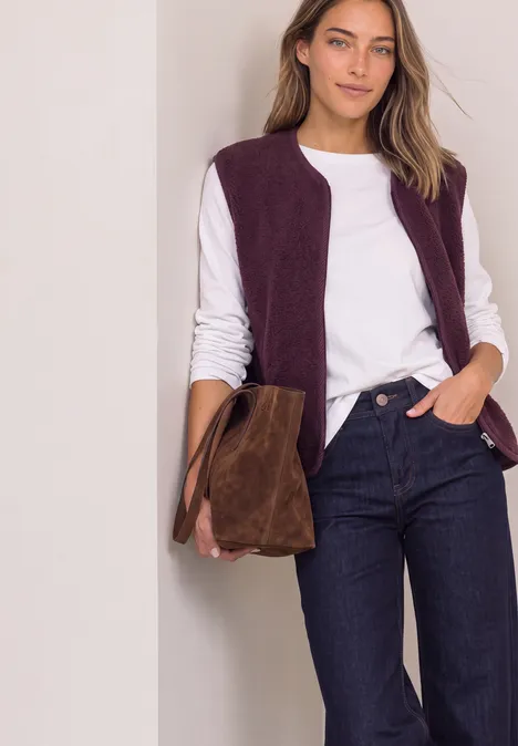 Vrouw met wit shirt, paarse bodywarmer en jeans, draagt een bruine suède tas voor een lichte achtergrond.
