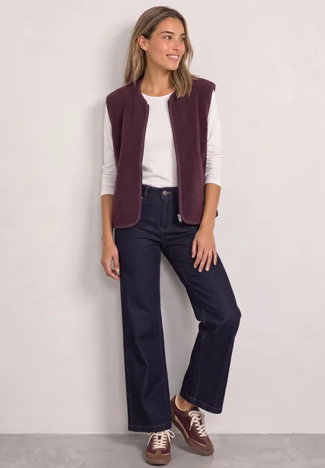 Vrolijke vrouw met wit shirt, paarse bodywarmer, wijde jeans en bruine schoenen voor een lichte achtergrond.
