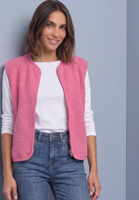 Een glimlachende vrouw in een roze mouwloos vest, wit T-shirt en spijkerbroek, voor een grijze achtergrond.