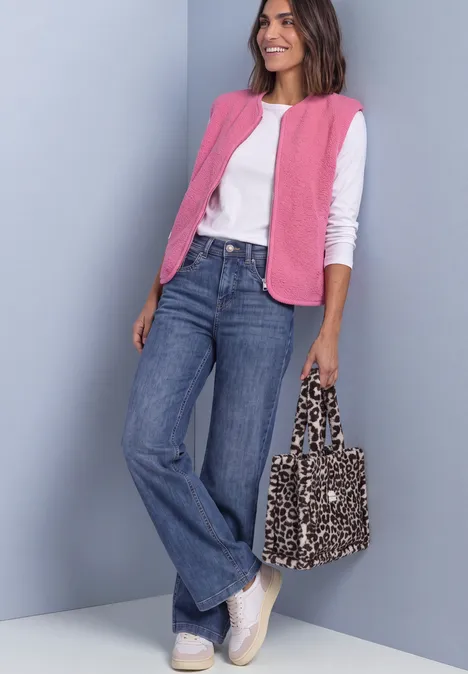 Vrouw in wit shirt, roze vest en wijde jeans, witte sneakers, glimlachend met een luipaardprint tas.
