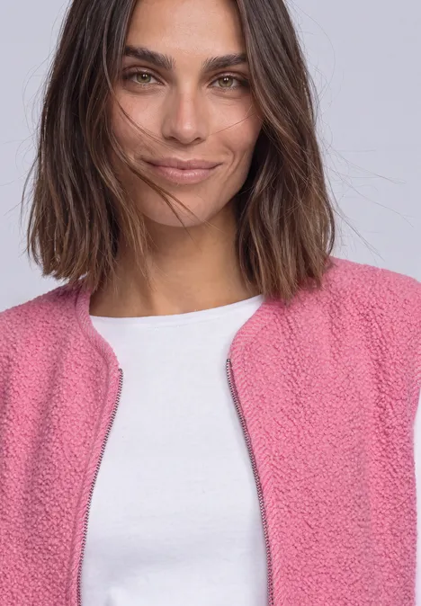 Een glimlachende vrouw met donker haar draagt een roze gebreid vest en een wit T-shirt.