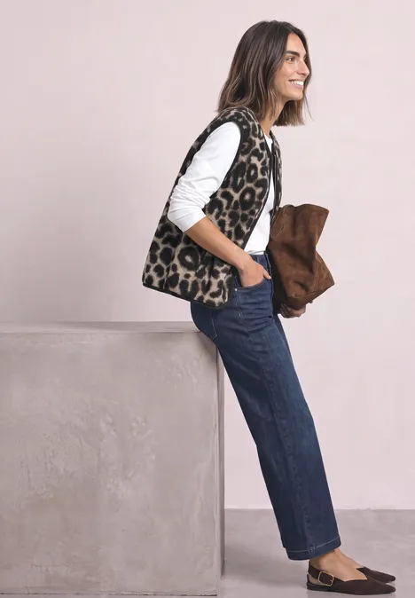 Gilet en peluche à fermeture zip et détails en velours côtelé Black Eine lächelnde Frau mit Leopardenmuster-Weste, weißem Shirt und Jeans lehnt an einem Block und hält eine braune Jacke.