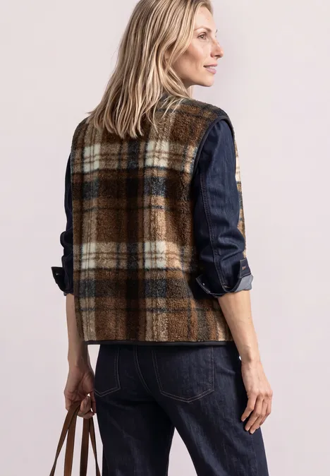 Gilet en peluche à fermeture zip et détails en velours côtelé Obsidian Blue Frau in karierter Wollweste und Jeanshemd, mit Lederhandtasche, seitlich stehend vor hellem Hintergrund.