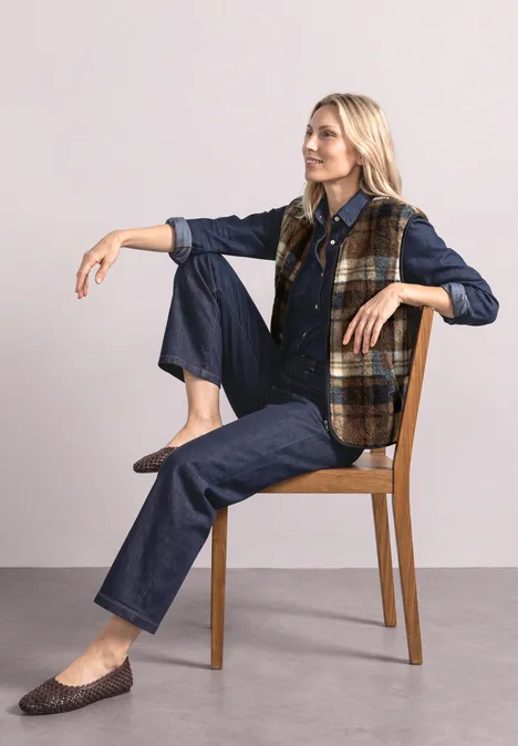 Gilet en peluche à fermeture zip et détails en velours côtelé Obsidian Blue Blonde Frau in Jeans, kariertem Weste und Loafers sitzt entspannt auf einem Stuhl.
