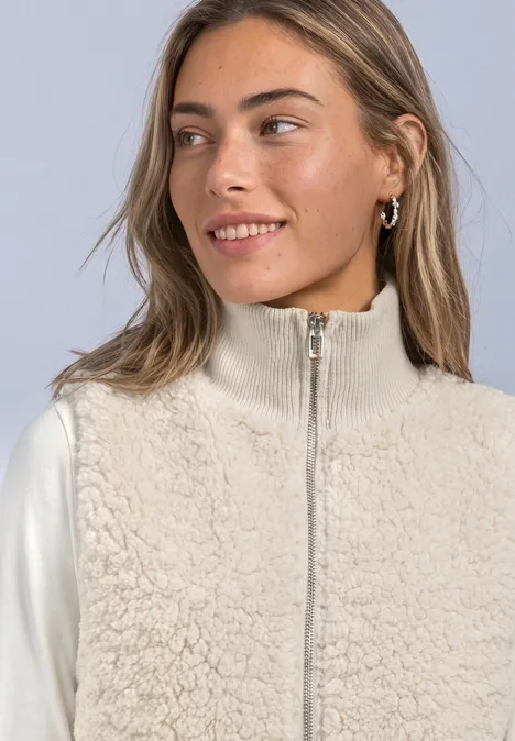 Junge Frau mit beiger Teddyfleece-Weste und weißem Langarmshirt, lächelnd nach rechts blickend.