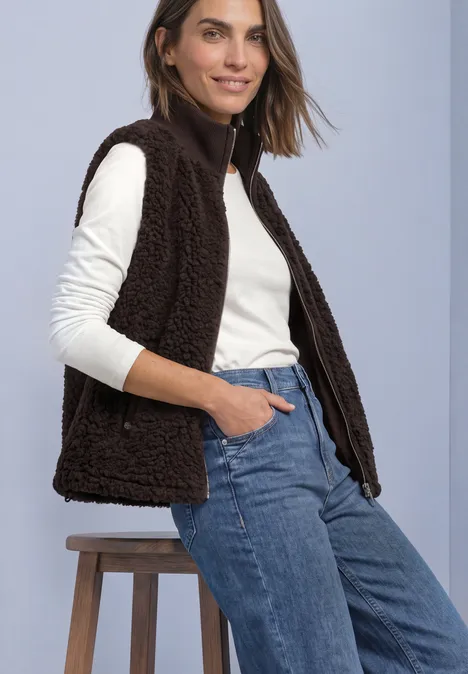 Frau mit weißem Longsleeve, flauschiger dunkelbrauner Weste und blauen Jeans, sitzt lässig auf einem Hocker.
