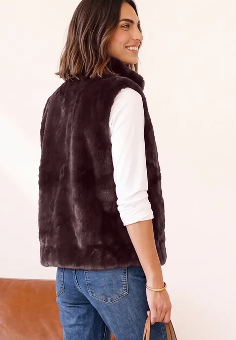 Vrouw in profiel met zwart faux bont gilet, witte longsleeve en jeans, glimlachend.