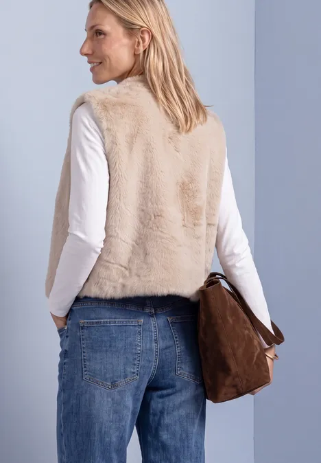 Frau von hinten in weicher, beiger Fake-Fur-Weste, weißer Bluse, blauen Jeans und brauner Tasche