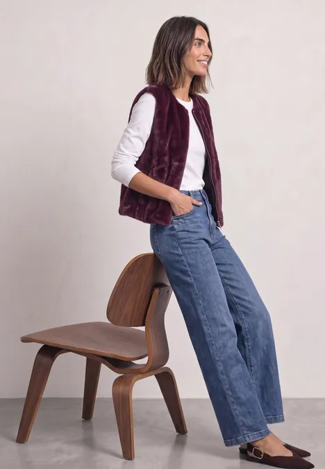 Frau in weißem Langarmshirt, weinroter Weste und weitem Jeans sitzt lächelnd an einem Holzstuhl.