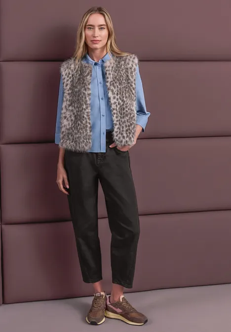 Eine Frau trägt ein graues Kunstfellweste über einer hellblauen Bluse, schwarze Hose und braune Sneaker.