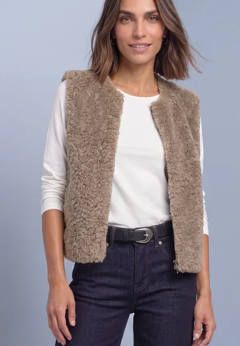 Frau trägt weiße Langarmshirt, beigen flauschigen Ärmellosen Weste und dunkle Jeans mit Gürtel vor grauem Hintergrund.