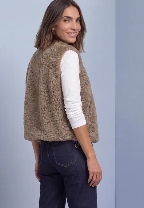 Eine Frau zeigt lächelnd von der Seite eine kamelfarbene Teddy-Weste über weißem Langarmshirt.
