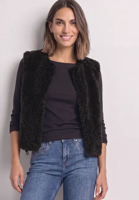 Vrouw met blauwe spijkerbroek, zwart shirt met lange mouwen en zwarte faux-fur gilet, glimlacht.