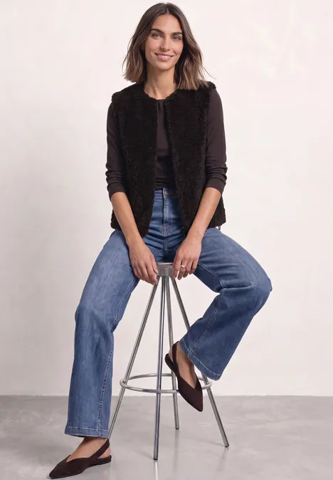 Vrouw zit lachend op barkruk, zwart jasje, jeans en bruine ballerina’s voor lichte achtergrond.