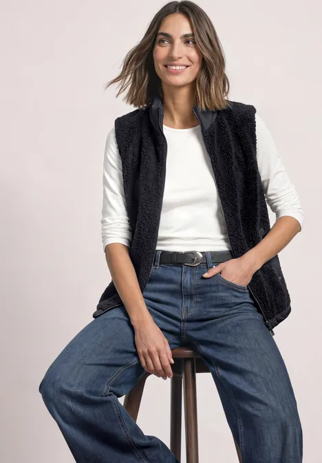 Vrouw zit op kruk, draagt zwarte teddy-vest, wit shirt en wijde jeans.