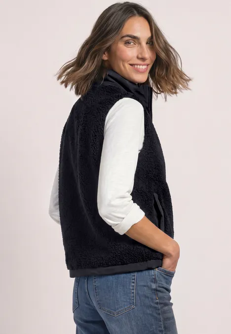 Een vrouw draagt een zwarte fleecebodywarmer over een witte longsleeve en spijkerbroek, kijkt schuin naar de camera.