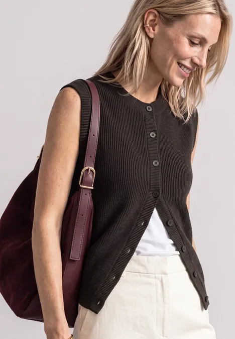 Eine lächelnde Frau trägt eine schwarze ärmellose Weste, ein weißes Top, einen hellen Rock und eine bordeauxfarbene Tasche.