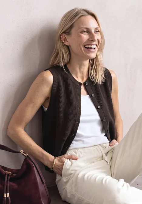 Eine lächelnde blonde Frau sitzt vor heller Wand, trägt weißes Top, braune Weste, helle Hose mit dunkelroter Tasche.