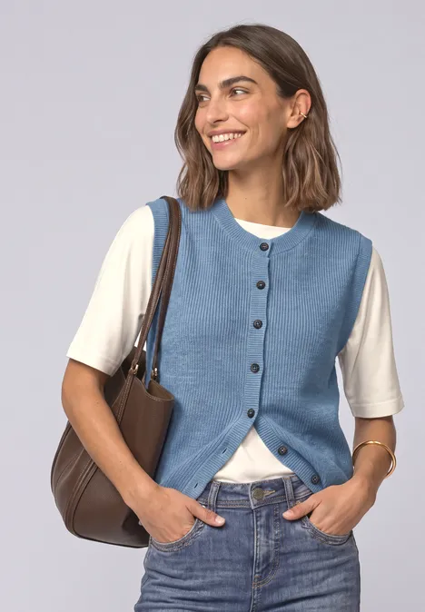 Frau mit blauer Weste, weißem Shirt, Jeans und brauner Tasche lächelnd in die Kamera.