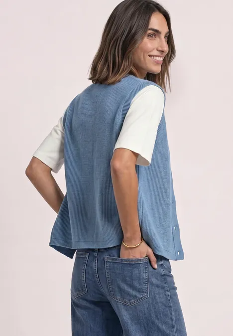 Eine lächelnde Frau trägt über einem hellen T-Shirt einen blauen, ärmellosen Pullunder und Jeans.