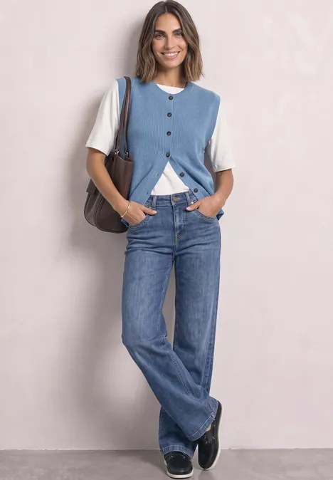 Frau mit blauem Strickweste, weißem T-Shirt, Jeans und Tasche vor hellem Hintergrund.