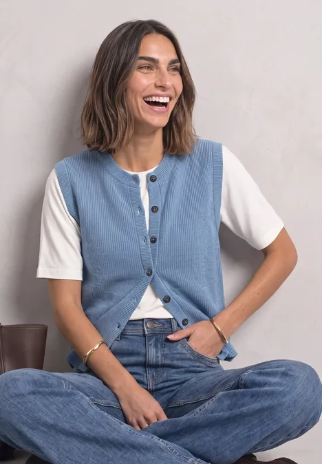 Eine lächelnde Frau sitzt im Schneidersitz, trägt eine weiße Bluse unter einer hellblauen Weste und blaue Jeans.