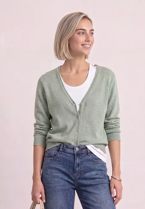 Junge Frau mit kurzem blondem Haar trägt hellgrüne Strickjacke, weißes Shirt und Jeans, sieht fröhlich aus.