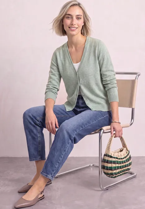 Cadigan mit V-Neck und Knopfleiste eucalyptus green mel. Frau in grünem Cardigan, Jeans und beigen Flats sitzt auf einem Stuhl und hält eine gewebte Tasche.
