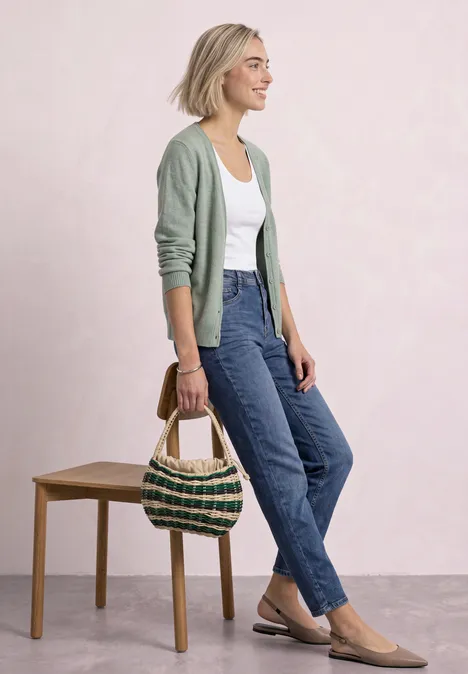 Cadigan mit V-Neck und Knopfleiste eucalyptus green mel. Frau mit grüner Strickjacke, weißem Top, blauen Jeans und Basttasche vor hellem Hintergrund.