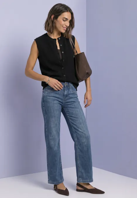 Strickweste mit Rundhals und Knopfleiste Black Frau mit schicker schwarzer Bluse, geraden Jeans, spitzen Schuhen und Lederhandtasche vor lila Hintergrund.