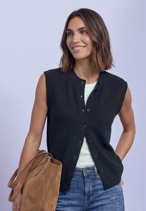 Frau trägt ärmellose schwarze Weste über weißem Top und hält eine braune Tasche, lächelnd.