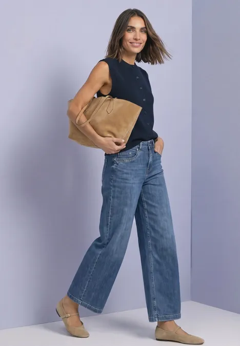 Frau in schwarzem ärmellosen Top, weiter blauer Jeans, beigen Slippern und großer brauner Tasche vor hellem Hintergrund.