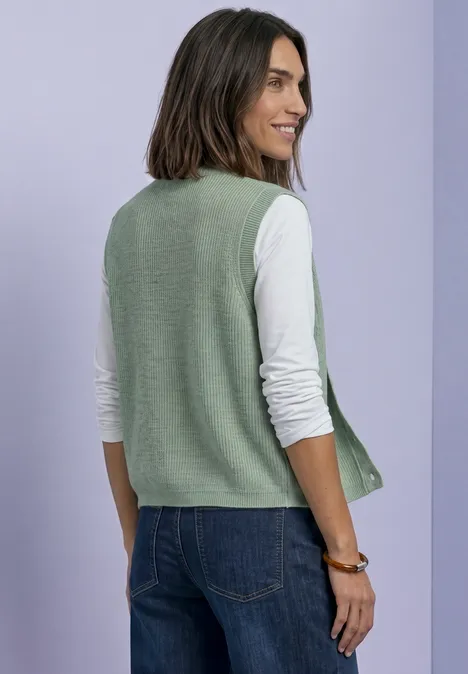 Eine Frau trägt eine grüne Strickweste über weißem Langarmshirt und Jeans vor lila Hintergrund.