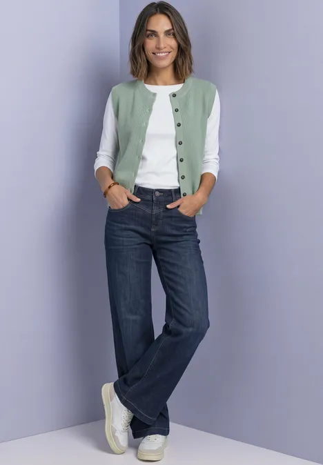 Frau mit grün-weißer Strickjacke, weißem Shirt, blauen Jeans und weißen Sneakern vor fliederfarbenem Hintergrund.