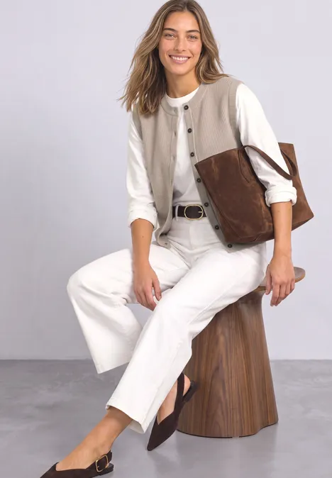 Eine Frau sitzt auf einem Hocker, trägt weiße Hose, weiße Bluse und braune Accessoires, lächelt freundlich.