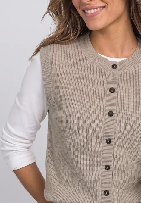 Frau im Halbprofil, lächelnd, trägt beige gerippte Strickweste mit Knöpfen über weißem Langarmshirt
